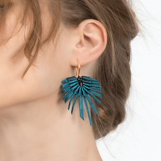 A white woman with blonde is wearing a pair of fan shaped earrings made with strands of teal textiles. The photograph shows a detail of her ear and neck.